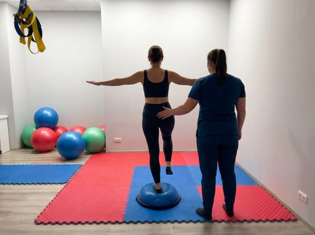 Pacjentka od tyłu stoi na jednej nodze na niebieskiej półkuli równoważnej, fizjoterapeutka koryguje ustawienie biodra i kolana — sala ćwiczeń CRMed